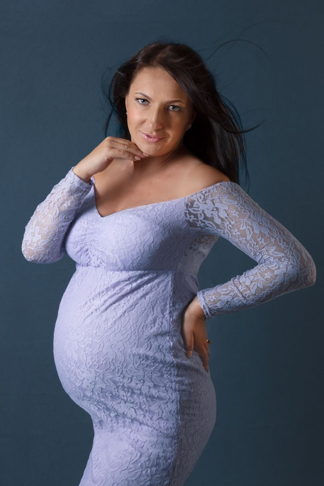 Studio maternity session of a pregnant woman wearing a light blue lace dress. The woman has long dark hair, she is looking straight into the camera smiling. It is a 3/4 portrait
