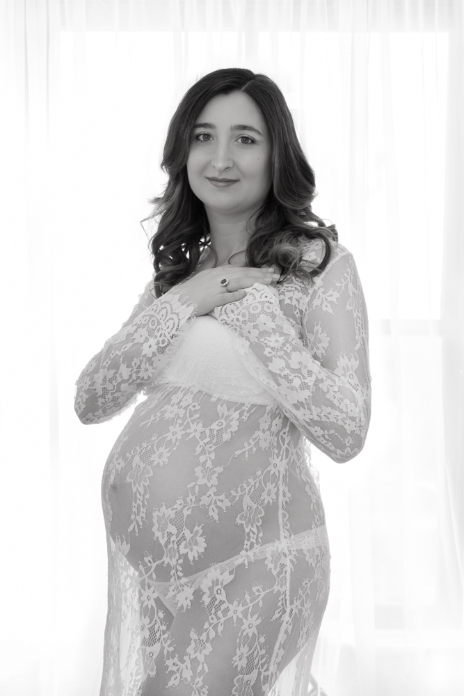 Fine art black and white image. Studio maternity session of a pregnant woman wearing a white lace dress. The woman has long dark hair, she is looking straight into the camera smiling. It is a 3/4 portrait