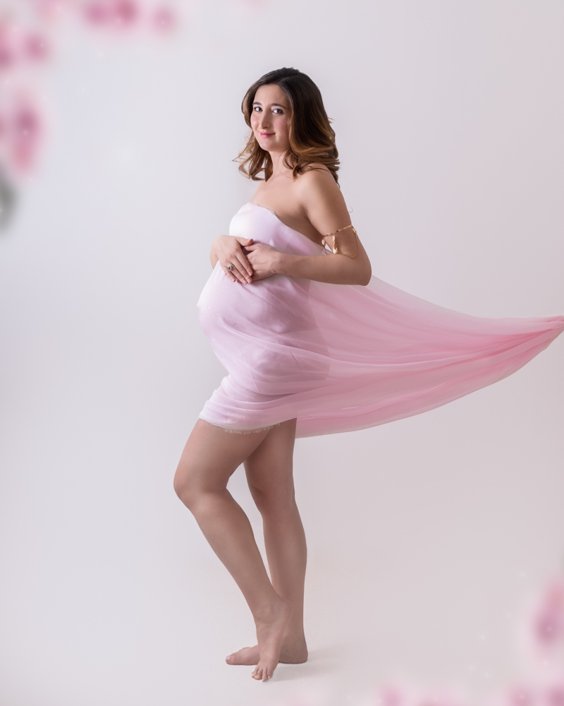 Studio maternity session of a pregnant woman with a pink chiffon fabric around her body. She is standing in front of a white background, profile posed but facing the camera. She is touching her belly with both hands and softly smiling.
