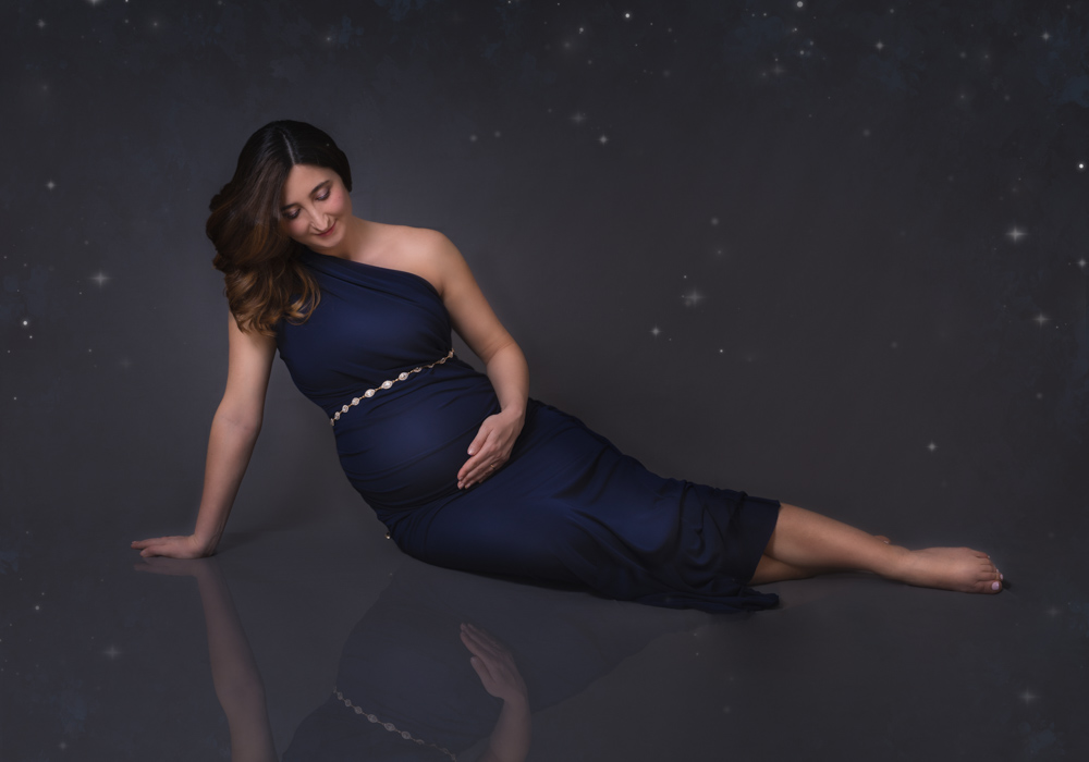 Fine art image. Studio maternity session of a pregnant woman wearing a blue long dress with a sparkling golden belt on top of her belly. She is sitting on her right handside with her legs slightly bent one on top of each other facing the other side. She is looking down towards her belly and with her left hand she is touching her belly. She is gently smile. She has long dark hair. The background is dark with some sparkles on it. A small part of her image is reflecting on the floor.