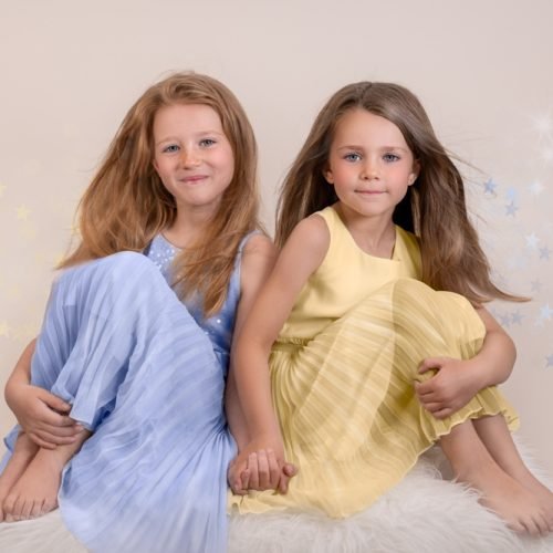 studio portrait of 2 young sisters. They are holding hands looking staright in to the camera. One is wearing a light blue dress and the other a yellow dress.