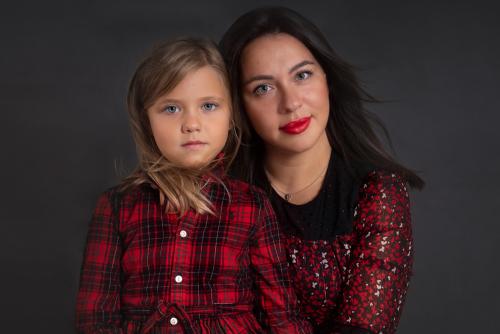 Mother and daughter wearing red and black outfit. It is shoulder portrait. The young girl has long blond hair, the mum has long dark hair. The girl is slightly in front of her mum. They are both looking at the camera. Image taken in studio in front of a grey background.