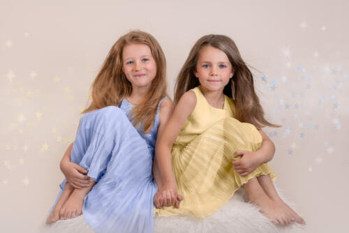 Portrait of two young sisters, wearing the same kind of dress but one is yellow and the other light blue. They are sitting on a white box, knees up and holding hands. 