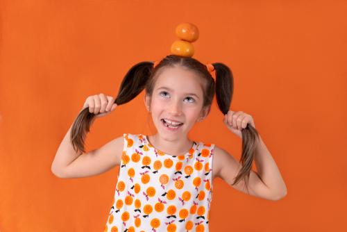 Studio portrait of a young girl in front of an orange background. She is holding her pig tails and looking up to two mandarins that are placed on top of her head
