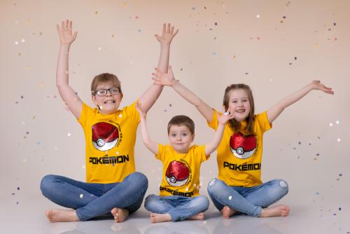 Photo of three young siblings (2 boys and a girl) wearing the same yellow top and blue jeans. Hands up after throwing confetti that are following on top of them. they are sitting on the floor and laughing.