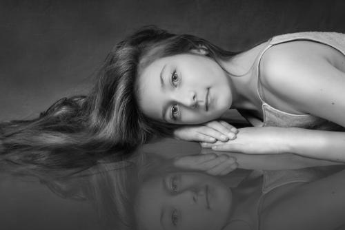 Young girl portrait in black white. She is laying on one side on the floor. Her hands, one on top of each other, are under her face. It is cropped to the shoulder. She is looking straight in to the camera.